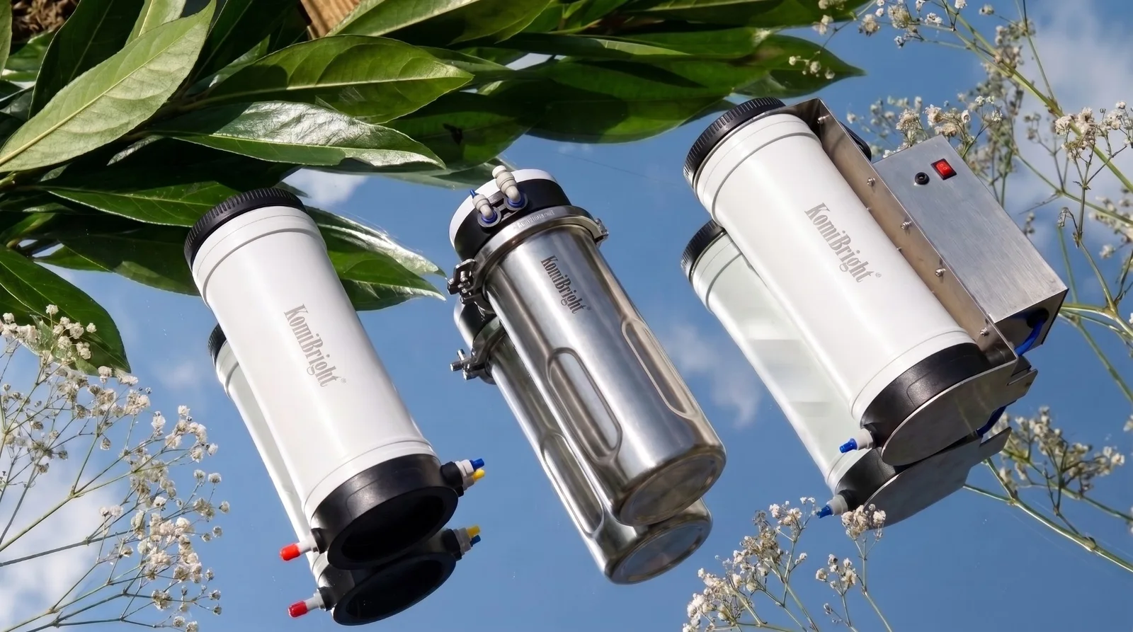 Three KomiBright systems photographed from below against a blue sky, among leaves and small white flowers.